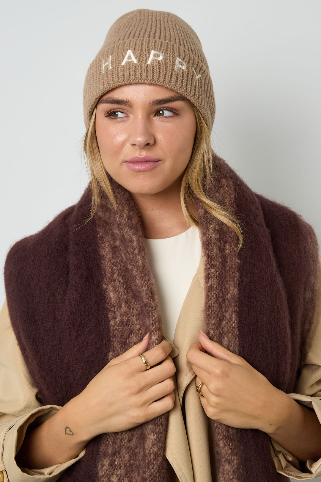 Model met khaki Happy muts en sjaal, close-up van de gebreide wintermuts met witte tekst.
