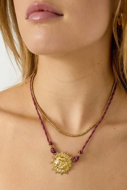 Close-up van korte ketting met zonhanger – wijnrood koord en goudkleurige zonhanger